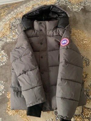 Grå dunjacka från Canada Goose - Säljer en grå dunjacka från Canada Goose med huva och hög krage. Jackan har ett klassiskt märke på ärmen, stora fickor framtill och stängs med både dragkedja och knappar. Perfekt för kalla vinterdagar och riktigt varm tack vare dunfyllningen.