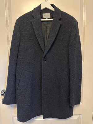 Velour Rock - Stilren mörkgrå rock från Velour med klassisk krage och två stora framfickor. Kappan är tillverkad i ett tjockt ullmaterial med diskret struktur och knappstängning framtill. Perfekt för kyliga dagar och ger en clean look.