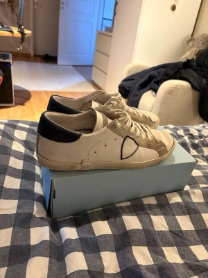Vita låga sneakers från Philippe Model - Säljer ett par vita låga sneakers från Philippe Model med beige detaljer och svart hälparti. Skorna har klassisk snörning, rund tå och är tillverkade i läder med mockadetaljer. Perfekta för en clean och stilren look.