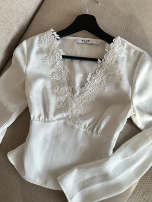 Vit blus med spetsdetaljer från NA-KD - Supersnygg vit blus från NA-KD med v-ringning och broderade spetsdetaljer runt halsen.💗💗 Bl. Materialet är glansigt, perfekt för att lyfta vilken outfit som helst. Storlek 32