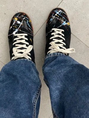Maison Margiela sneakers med färgstänk - Unika svarta sneakers från Maison Margiela med vita, blå och gula färgstänk. Skorna har vita snören och klassisk gummisula. Perfekt för dig som vill sticka ut med en kreativ och konstnärlig vibe. Materialet är läder och sulan är i gummi.
