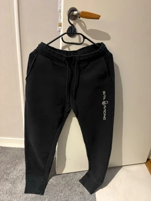 Svarta mjukisbyxor från Sail Racing - Svarta mjukisbyxor från Sail Racing med logga längs ena benet. Byxorna har dragsko i midjan, muddar vid bensluten och två sidofickor. Tillverkade i mjukt material som känns skönt mot huden. Perfekta för chill eller träning.