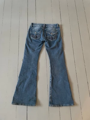 Blå lågmidjade bootcut jeans från Gina Tricot - Säljer ett par blå bootcut jeans med slitningar på knäna och ljusa tvättade partier. Jeansen har detaljerade bakfickor med lock och knappar. Perfekta för en avslappnad och trendig look. Säljer byxorna då jag tyvärr har växt ur dem. De har väldigt små slitage längst ner men syns om man inte tittar noga. 