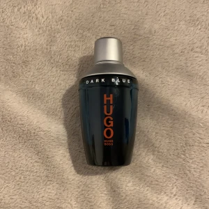 Hugo Boss Dark Blue parfym - En stilren parfymflaska från Hugo Boss, modell Dark Blue. Flaskan är mörkblå med silverfärgat lock och har tydlig orange text på framsidan. Designen är modern och flaskan är tillverkad i glas med metallock. Perfekt för dig som gillar klassiska dofter med en twist.