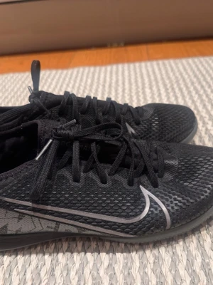 Svarta Nike fotbollsskor inomhus - Säljer ett par svarta Nike fotbollsskor för inomhusbruk med låg profil och slitstark gummisula. Skorna har ett ventilerande meshmaterial och klassisk snörning. Swoosh-logga i silver på sidan och undersidan. Perfekta för futsal eller inomhusträning.
