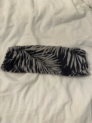 Svart och grå bandeau bikiniöverdel - Säljer en bandeau bikiniöverdel i svart och grått med ett stilrent palmbladsmönster. Modellen är axelbandslös och har resår i kanterna för att sitta på plats. Perfekt för dig som vill ha en enkel men snygg bikiniöverdel till stranden.