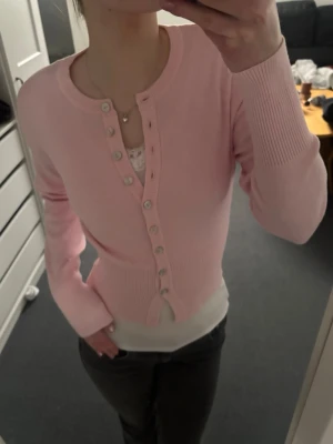 Ljusrosa cardigan från Gina Tricot - Söt ljusrosa cardigan från Gina Tricot med rund halsringning och långa ärmar. Tröjan har ribbade muddar och knappar framtill i pärlemor-look. Perfekt att styla över ett linne eller t-shirt för en fräsch och trendig look.