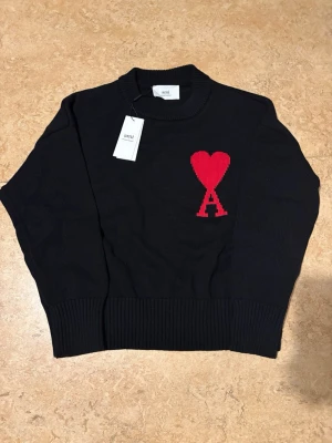 Svart stickad tröja från Ami Paris. Storlek: S - Svart stickad tröja från Ami Paris med röd hjärtlogga och bokstaven A på bröstet. Tröjan har rund halsringning, ribbade muddar och är gjord i mjukt material. Perfekt för dig som gillar stilrena detaljer och streetwear-vibe.