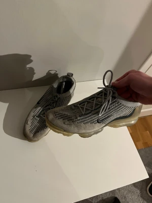Nike Air Vapormax grå sneakers - ÄKTA Nike Air Vapormax sneakers i grått med stickad ovandel och transparent sula. Skorna har en låg profil, synlig Air-dämpning och svarta detaljer på sidorna samt blå Nike-logga på hälen. Perfekta för dig som gillar sportig och modern stil. Spräckt bubbla som man ser på sista bilden men känns knappt när man går så funkar defenitivt o använda som vanligt