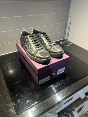 Snygga Baldessarini "Gary" Sneakers i Svart Läder – Nyskick! - Säljer ett par exklusiva och stilrena sneakers från tyska lyxmärket Baldessarini, modell "Gary". Skorna är tillverkade i högkvalitativt svart glatt läder med en subtil men elegant logotypprägling över hela ovandelen, vilket ger dem en unik karaktär. Dessa sneakers kombinerar en sportig silhuett med en dressad känsla, vilket gör att de passar lika bra till kostymbyxor som till jeans.