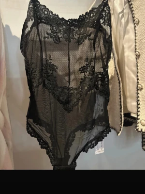 Svart spetsbody med blommönster - Snygg svart body i transparent mesh med detaljerad spets och blommönster. Djup urringning och dekorativa kanter ger en feminin look. Det Perfekt för dig som vill ha något extra under kläderna. Materialet känns lätt och följsamt.