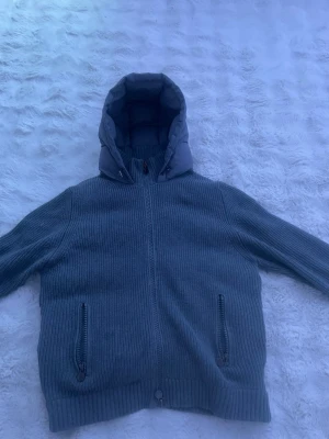 Grå pufferjacka med stickade ärmar - Snygg grå pufferjacka med stickade ärmar och ribbade muddar. Jackan har en vadderad huva och baksida, samt två framfickor med dragkedja. Perfekt för kalla vinterdagar och ger en cool, avslappnad look. Pris går att diskutera 