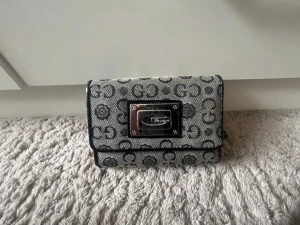 Grå mönstrad plånbok från Guess - Snygg grå plånbok från Guess med svart logomönster och blommor över hela ytan. Plånboken har en silverfärgad metallplatta med Guess-logga på framsidan och svarta kanter. Tillverkad i textil med detaljer i konstläder. Kan diskutera pris villig att sänka💓