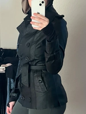Vintage Svart trenchcoat i skinnlook - Snygg svart trenchcoat med dubbelknäppning och bälte i midjan. Jackan har klassisk krage, axelklaffar och dekorativa knappar vid ärmsluten. Materialet ser ut som skinn och ger en cool, stilren vibe. Perfekt för dig som vill ha en edgy och tidlös jacka.