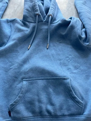 Blå hoodie från Superdry - Snygg blå hoodie från SuperDry med broderad logga på bröstet och klassisk känguruficka framtill. Hoodien har dragsko i huvan och ribbade muddar. Perfekt för en chill och avslappnad stil. Tillverkad i mjukt bomullsmaterial. Tröjan är superbra skick utan hål eller fläckar. Priset kan diskuteras!