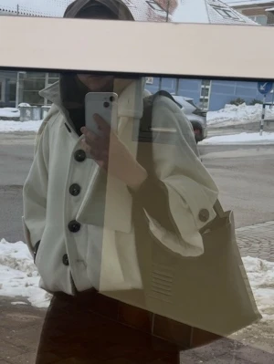 Vit trenchcoat med stora knappar - Stilren vit trenchcoat med stora svarta knappar och bred krage. Jackan har bälte i midjan och dekorativa knappar vid ärmsluten. Materialet ser ut att vara ullblandning, vilket ger en mjuk och varm känsla. Perfekt för dig som vill ha en klassisk men trendig look.