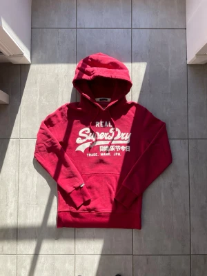 Röd hoodie från Superdry - Snygg röd hoodie från Superdry med vit logotyp och japanska tecken på bröstet. Klassisk huva med dragsko och ribbade muddar. Perfekt för dig som gillar streetwear och vill ha något bekvämt och stilrent. Materialet är mjukt och känns riktigt skönt. Vid frågor eller funderingar skriv gärna!