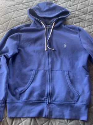 Blå hoodie från Polo Ralph Lauren - Blå zip-hoodie från Polo Ralph Lauren med vit broderad logga på bröstet. Hoodien har dragsko i huvan, två stora fickor framtill och ribbade muddar. Tillverkad i mjuk bomull för skön känsla och avslappnad stil.