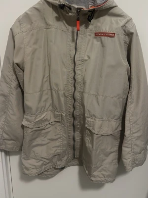 Beige vindjacka från KappAhl Boys - Stilren beige vindjacka från KappAhl Boys med huva, dragkedja och två stora fickor framtill. Jackan har en röd detalj vid dragkedjan och Street Zone-märke på bröstet. Perfekt för blåsiga dagar och enkel att matcha med olika outfits.