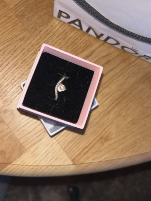 Silverring med hjärta från Pandora - Snygg silverring från Pandora med ett glittrande hjärta i mitten och små stenar längs bandet. Ringen har en elegant och modern design som verkligen sticker ut. Perfekt för dig som gillar stilrena smycken med lite extra bling.