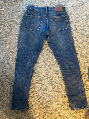 Blå sopade Levi's jeans - Ett par klassiska blå jeans från Levi's med raka ben och två bakfickor med den ikoniska sömmen. Jeansen har en brun läderpatch i midjan och en röd Levi's-tagg på fickan. Tillverkade i slitstarkt denimtyg, perfekta för en avslappnad look.