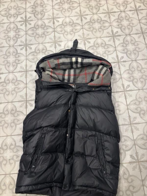 Svart burberry dunväst med huva och rutigt foder - Snygg svart dunväst med stor huva och dragkedja framtill. Insidan av huvan har ett rutigt foder i grått, rött och vitt. Västen har två dragkedjefickor och breda kanaler för extra värme. Perfekt för kalla dagar och enkel att matcha.