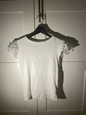 Vit topp med spetsärmar - Söt vit topp med korta puffärmar i broderad spets och rund halsringning. Kroppen är i mjuk ribbad bomull som ger en skön känsla och snygg passform. Perfekt för dig som gillar romantiska detaljer och stilren look.