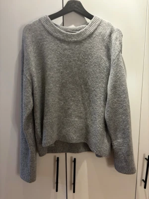 Oversiced Grå stickad tröja med rund hals - Mysig grå stickad tröja med rund halsringning och breda ribbade muddar vid ärmslut och nederkant. Tröjan har en avslappnad passform och är perfekt för lager på lager. Passar dig som gillar enkel och stilren design.