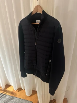 Moncler cardigan  - Snygg svart pufferjacka från Moncler med quiltad front, tvåvägsdragkedja och diskret Moncler-logga på ärmen. Jackan har två dragkedjeförsedda fickor och ribbade muddar. Perfekt för kalla vinterdagar och ger en clean, modern look.