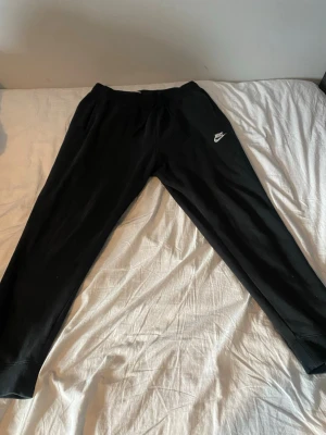 Svarta Nike mjukisbyxor - Säljer ett par svarta mjukisbyxor från Nike med klassisk vit logga på vänster lår. Byxorna har en avslappnad passform, resår i midjan och muddar vid bensluten. Perfekta för chill dagar eller när du vill ha det bekvämt.