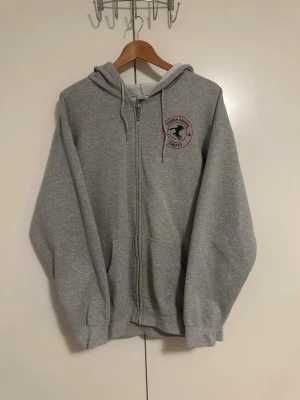 Grå Vintage Champion hoodie - Grå zip-hoodie med Parma Animal Shelter-logga på bröstet och stor logga på ryggen. Klassisk modell med huva och dragsko, samt fickor framtill. Tillverkad i mjukt sweatshirtmaterial, perfekt för chill dagar eller promenader.