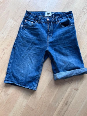 Blå jeansshorts med uppvik - Snygga blå jeansshorts från 157 med klassisk femficksdesign och uppvikta benslut. Shortsen har normal passform och är tillverkade i slitstark bomull. Perfekta för varma dagar och enkel att matcha med olika stilar.