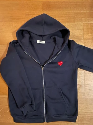 Mörkblå hoodie från Comme des Garçons - Säljer en mörkblå hoodie med dragkedja från Comme des Garçons PLAY. Hoodien har en huva, två fickor framtill och det ikoniska röda hjärtat med ögon broderat på bröstet. Perfekt för en avslappnad och stilren look.