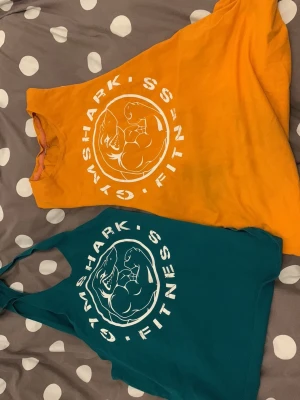 Gymshark linnen i orange och petrol - Två snygga linnen från Gymshark, ett i stark orange och ett i petrolblått. Båda har stort vitt tryck med Gymshark-logga framtill och är ärmlösa med djupa ärmhål, perfekta för gymmet eller träning. Ena medium andra lite större som L. Og David Laid gym era gymshark 