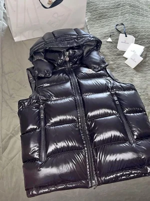 Svart bormes dunväst från Moncler - Snygg svart dunväst från Moncler med glansig finish och vadderad design. Västen har två dragkedjefickor framtill, hög krage och avtagbar huva. Insidan har Moncler-logga och en unik serieteckning. Perfekt för lager på lager och streetstyle. Passar på 165-180 cm 