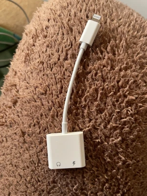 Apple Lightning till dubbla adaptrar - Praktisk vit adapter från Apple med Lightning-kontakt och två portar för laddning och hörlurar. Perfekt för dig som vill ladda mobilen och lyssna på musik samtidigt. Tillverkad i tålig plast med smidig och kompakt design.