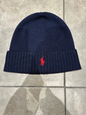 Mörkblå mössa från Polo Ralph Lauren - Snygg mörkblå ribbstickad mössa från Polo Ralph Lauren med klassisk röd broderad logga framtill. Mössan har uppvikt kant och är tillverkad i mjukt stickat material som håller dig varm under kalla dagar.