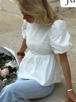 Vit blus med puffärm och rosett - Supersöt vit blus med puffärmar och öppen rygg som knyts med både band och stor rosett. Blusen har en lätt utsvängd passform och är perfekt för dig som gillar romantiska detaljer. Tillverkad i ett mjukt bomullsliknande material.