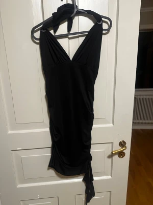 Svart kortklänning med knyt - Snygg svart kortklänning med omlottdesign och knyt i nacken. Klänningen har djup v-ringning och draperad effekt framtill 