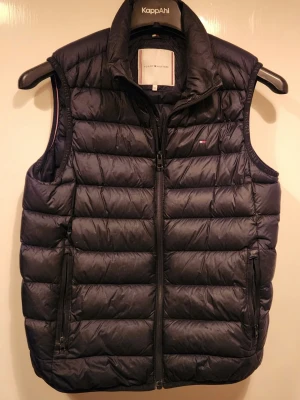 Mörkblå dunväst från Tommy Hilfiger - Snygg mörkblå dunväst från Tommy Hilfiger med quiltad design och hög krage. Västen har två dragkedjefickor framtill och diskret logga på bröstet. Perfekt att slänga över en hoodie eller tröja för extra värme. Klassisk och stilren look.