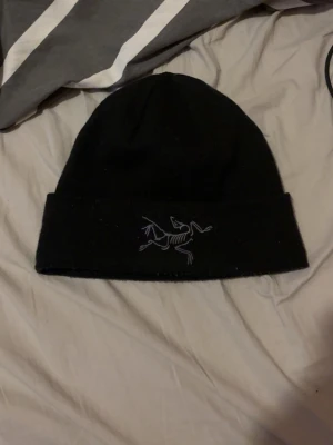 Svart mössa från Arc'teryx - Säljer en svart mössa från Arc'teryx med den ikoniska vita loggan broderad framtill. Klassisk modell med uppvikt kant, perfekt för kalla dagar. Materialet känns mjukt och värmande, passar till de flesta outfits.