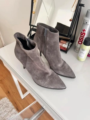 Gråa spetsiga stövletter i mocka - Säljer ett par snygga gråa stövletter i mocka med spetsig tå och smal klack. Dragkedja på insidan för enkel på- och avtagning. Perfekta för dig som vill ha en stilren och trendig look till hösten eller vintern. Använda en gång.