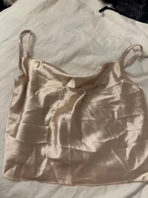 Champagnefärgad satin linnetopp - Snygg champagnefärgad linnetopp i satin med tunna axelband och slät finish. Perfekt att styla med jeans eller kjol för en trendig look. Enkel och stilren design som känns lyxig och modern.