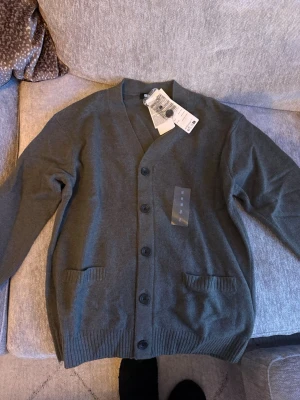 Mörkgrön kofta med knappar - Stilren mörkgrå kofta med v-ringning och knäppning framtill. Koftan har två fickor fram och ribbade muddar vid ärmslut och nederkant. Perfekt för lager-på-lager och enkel att matcha med olika outfits.