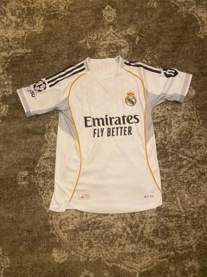 Real Madrid matchtröja Vini Jr. - Snygg vit Real Madrid matchtröja .med svarta och gula detaljer. Tryck med 'Emirates Fly Better' på framsidan och 'VINI JR. 7' på ryggen. Tröjan har klubbmärke, Champions League-märke på ärmen och är tillverkad i ett lätt funktionsmaterial. Storlek XS och kan passa M