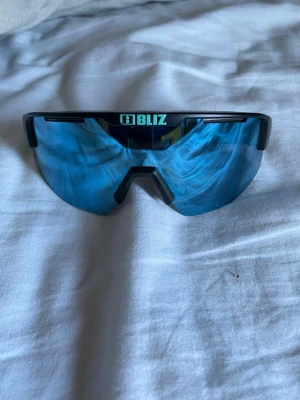 Blå sportiga solglasögon från Bliz - Säljer ett par sportiga solglasögon från Bliz med blå spegelglas och svart båge. Glasögonen har en modern, kantig form och är perfekta för utomhusaktiviteter. Tillverkade i lätt syntetmaterial för maximal komfort. Fick glasögonen i julklapp men använda enstaka gånger, vet ej vart boxen är.