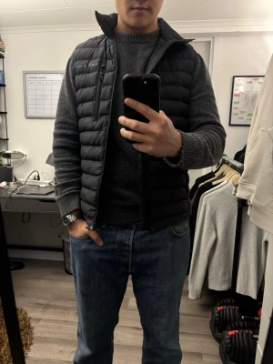 Svart dunväst från Peak Performance - Snygg svart dunväst från Peak Performance med hög krage och dragkedja framtill. Västen har tre fickor med dragkedja och quiltad design. Perfekt att slänga över en hoodie eller tröja för extra värme. Klassisk och stilren look som funkar året runt.