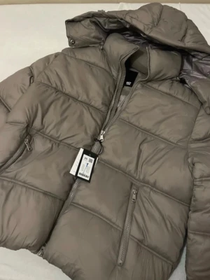 Brun/khakipufferjacka  - Säljer en brun pufferjacka från 157 med huva och dragkedja framtill. Jackan har två sidofickor med dragkedja och quiltad design. Materialet är glansigt och jackan är perfekt för kalla vinterdagar. Enkel och stilren modell som håller dig varm.