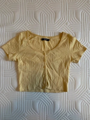 Gul croptop Bik Bok - Söt ljusgul croppad topp från Bik Bok med korta ärmar och rund halsringning. Toppen har knappar framtill och ett diskret mönster i tyget.
