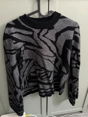 Svart och silvermönstrad stickad tröja - Stickad tröja i svart med silvrigt zebramönster. Tröjan har rund halsringning med ribbad kant och långa ärmar. Materialet ser glittrigt ut och ger en cool kontrast mellan svart och silver. Perfekt för dig som gillar att sticka ut med mönster.